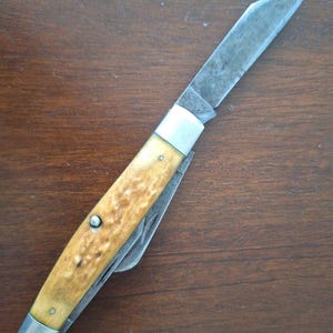 Puede incluir: Una navaja de bolsillo vintage con una hoja de acero inoxidable desgastada y un mango con un patr&oacute;n texturizado marr&oacute;n claro. La navaja tiene virolas plateadas y remaches. La hoja est&aacute; parcialmente abierta, revelando una hoja secundaria m&aacute;s peque&ntilde;a.