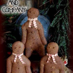 May include: Three gingerbread people dolls with red and white striped scarves. The dolls are made of brown fabric and have button eyes. The dolls are sitting on a red and white checkered fabric.