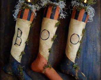 Puede incluir: Tres adornos de tela con forma de calcetín con rayas negras y naranjas, cada uno con una pequeña luz en el interior y una letra pintada que deletrea "BOO" sobre un fondo de madera oscura.