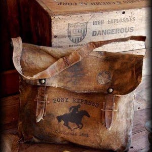 Może przedstawiać: Brązowa torba płócienna w stylu vintage z napisem "PONY EXPRESS" i sylwetką jeźdźca na koniu. Torba ma klapę z paskami skórzanymi i metalowymi okuciami. Widoczny jest również tekst "DAIL 1862". W tle znajduje się drewniana skrzynia.