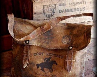 Op de afbeelding: Een bruine canvas tas in vintage stijl met de woorden "PONY EXPRESS" en een silhouet van een ruiter op een paard. De tas heeft een klepsluiting met leren riemen en metalen hardware. De tekst "DAIL 1862" is ook zichtbaar. Op de achtergrond staat een houten kist.
