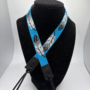 May include: A beaded camera strap featuring a vibrant blue background with a repeating pattern of white, gray, and black feathers. The strap is finished with black leather end tabs and thin black cord attachments for securing to a camera body.