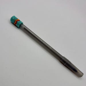 May include: A translucent dark gray pen featuring a decorative cap adorned with small, multi-colored glass beads. The beadwork displays a geometric pattern in shades of turquoise, orange, and red, covering the top portion of the writing instrument.