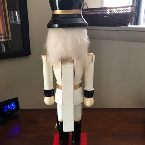 May include: A white wooden nutcracker with a black hat and gold accents. The nutcracker is standing on a red base.