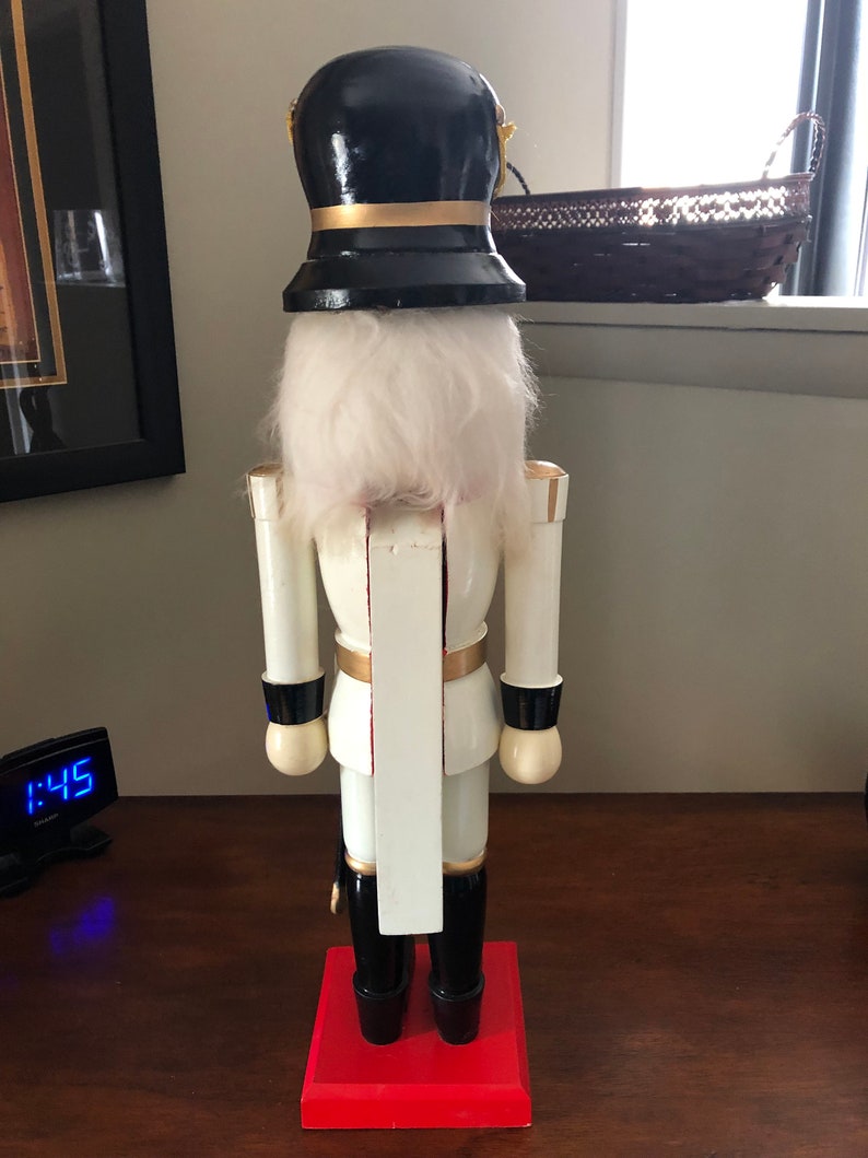 May include: A white wooden nutcracker with a black hat and gold accents. The nutcracker is standing on a red base.