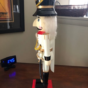May include: A white wooden nutcracker with a black hat, red and gold accents, and a black sword. The nutcracker is standing on a red base.