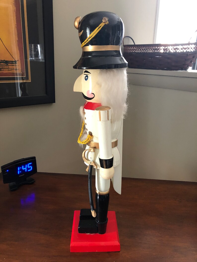 May include: A white wooden nutcracker with a black hat, red and gold accents, and a black sword. The nutcracker is standing on a red base.