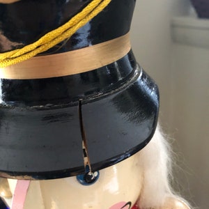 May include: A vintage wooden nutcracker with a black hat, gold trim, and a white beard. The nutcracker has a painted face with blue eyes and a pink mouth. The nutcracker is holding a wooden cup in its right hand.