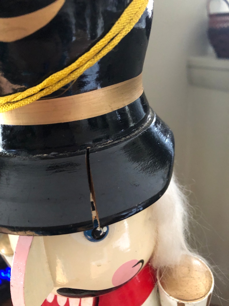 May include: A vintage wooden nutcracker with a black hat, gold trim, and a white beard. The nutcracker has a painted face with blue eyes and a pink mouth. The nutcracker is holding a wooden cup in its right hand.