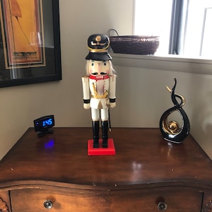 May include: A wooden nutcracker figurine with a white and gold uniform, black hat, and red base. The nutcracker is standing on a wooden table with a clock and a black and gold abstract sculpture.