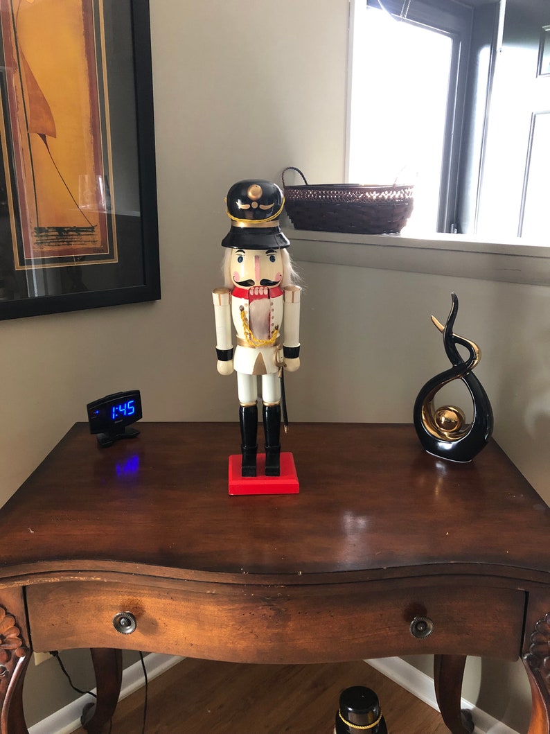 May include: A wooden nutcracker figurine with a white and gold uniform, black hat, and red base. The nutcracker is standing on a wooden table with a clock and a black and gold abstract sculpture.