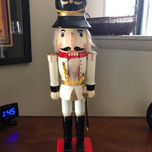 May include: A wooden nutcracker figurine dressed as a soldier. The nutcracker is wearing a black hat with gold trim, a white uniform with gold accents, and black boots. The nutcracker is holding a sword in its right hand.