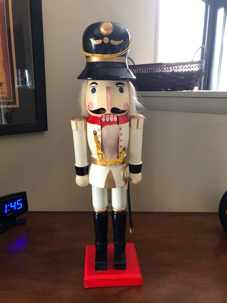 May include: A wooden nutcracker figurine dressed as a soldier. The nutcracker is wearing a black hat with gold trim, a white uniform with gold accents, and black boots. The nutcracker is holding a sword in its right hand.