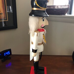 May include: A white wooden nutcracker with a black hat and gold trim. The nutcracker is standing on a red base.