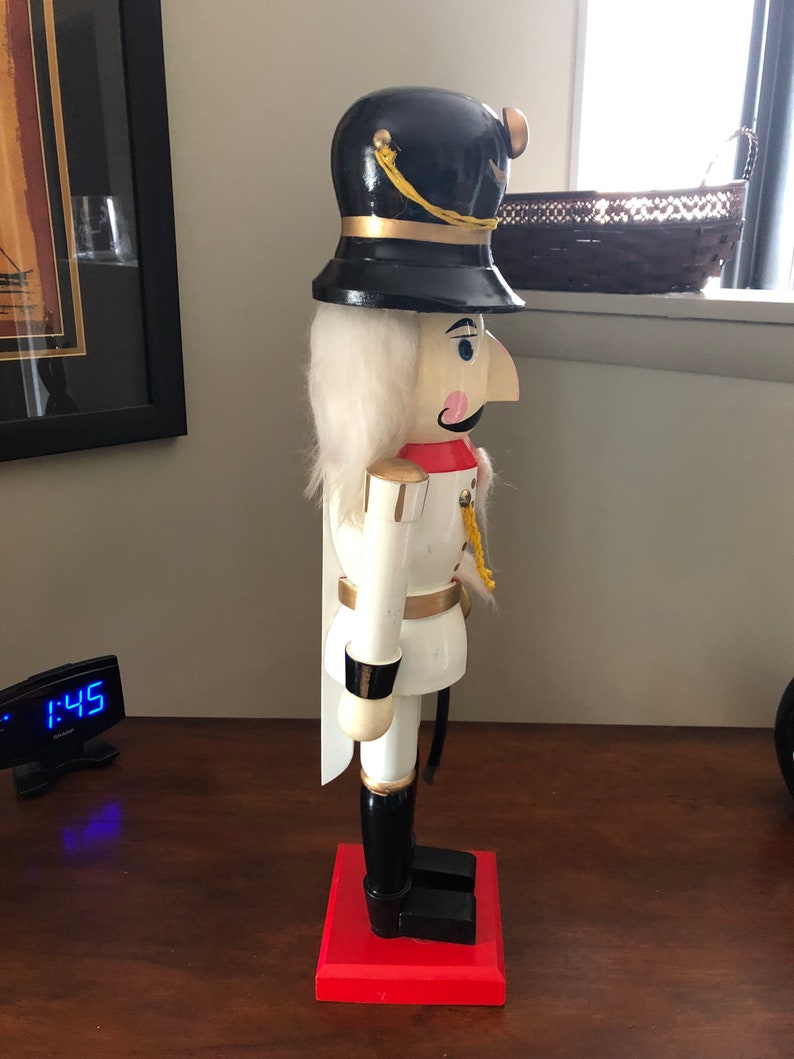 May include: A white wooden nutcracker with a black hat and gold trim. The nutcracker is standing on a red base.