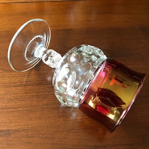 May include: A clear glass goblet with a textured, bubbled design and a red iridescent base. The goblet is on a wooden surface.
