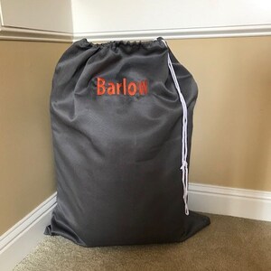 May include: A dark gray drawstring bag with the name "Barlow" embroidered in orange. The bag is made of a smooth fabric and has a white drawstring closure. It is sitting on a carpeted floor.