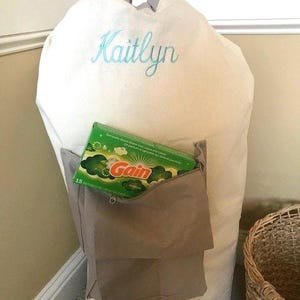 May include: A white canvas laundry hamper with a brown fabric pocket and a zipper. The hamper has the name "Kaitlyn" embroidered on the front in teal blue thread. A box of Gain laundry detergent is in the pocket.