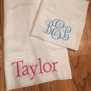 May include: Two white pillowcases with embroidered lettering. One pillowcase has the monogram "BEL" in blue thread. The other pillowcase has the name "Taylor" in pink thread.
