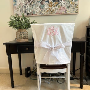May include: White chair cover with a pink monogram "LSA" embroidered on the back. The chair cover has a white satin ribbon tied in a bow at the back.