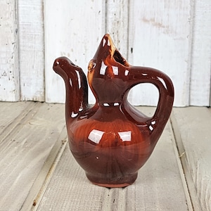 Può includere: Una teiera in ceramica marrone con un design unico. La teiera ha un corpo arrotondato e un beccuccio che si curva verso l'alto. Il manico è sul lato della teiera ed è anche arrotondato.