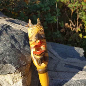 May include: A wooden hand-carved figurine of a face with a wide open mouth, painted in yellow, red, black, and white. The figurine is a whimsical and unique piece of folk art.