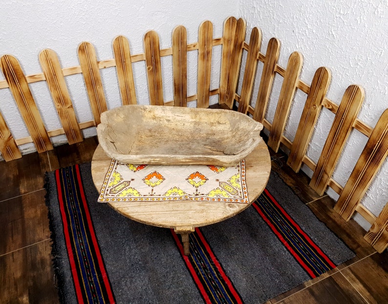 K&ouml;nnte beinhalten: Eine Holzsch&uuml;ssel steht auf einem runden Holztisch mit einer gemusterten Tischdecke. Der Tisch steht auf einem grauen und rot gestreiften Teppich. Ein Holzzaun umgibt den Tisch.