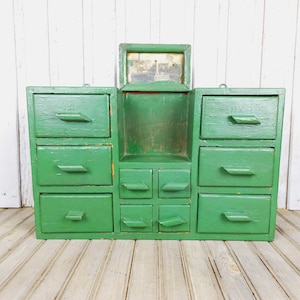 Könnte beinhalten: Ein antiker grüner Holzschrank mit mehreren Schubladen und einem kleinen offenen Regal. Der Schrank hat einen kleinen Bilderrahmen oben.