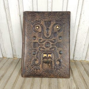May include: A brown leather-bound journal with an embossed design. The cover features an intricate pattern with gold and green accents. The journal is rectangular and appears to be vintage or antique.