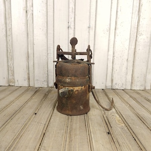 May include: A vintage, rusty metal lantern with a handle and hook. The lantern has a cylindrical body with a series of metal bands and a top with a screw mechanism. The overall color is brown with signs of age and wear.