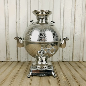 May include: A silver samovar with an ornate design, featuring a round body, a spout, and two handles. The samovar is sitting on a wooden surface.
