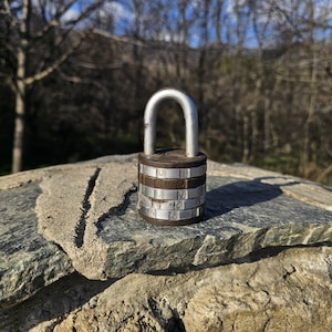 May include: A combination lock with a silver shackle and a dark brown and silver body. The lock is set on a stone surface with a blurred background of trees and sky. The lock's dials display numbers.