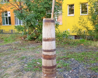 May include: A wooden barrel with metal bands, standing upright on a gravel surface. The barrel is made of light brown wood and has a natural finish. The metal bands are silver in color and are evenly spaced around the barrel.