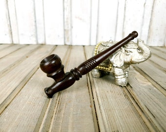 May include: A dark brown wooden smoking pipe with a round bowl and a long stem. The pipe is lying on a wooden surface next to a silver elephant figurine.