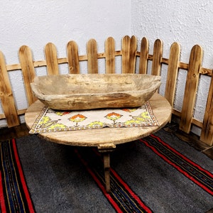 K&ouml;nnte beinhalten: Ein Holztisch mit runder Tischplatte und drei Beinen. Eine Holzsch&uuml;ssel steht auf einem gemusterten Tischtuch auf dem Tisch. Der Tisch steht vor einem Holzzaun.