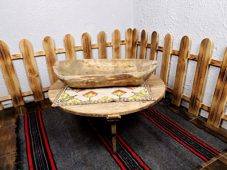 K&ouml;nnte beinhalten: Ein Holztisch mit runder Tischplatte und drei Beinen. Eine Holzsch&uuml;ssel steht auf einem gemusterten Tischtuch auf dem Tisch. Der Tisch steht vor einem Holzzaun.