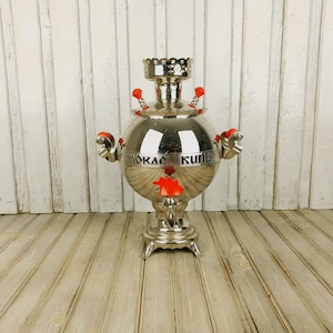 May include: A silver samovar with red accents and the inscription "Мокрая Кухня" on the side. The samovar is sitting on a wooden floor.