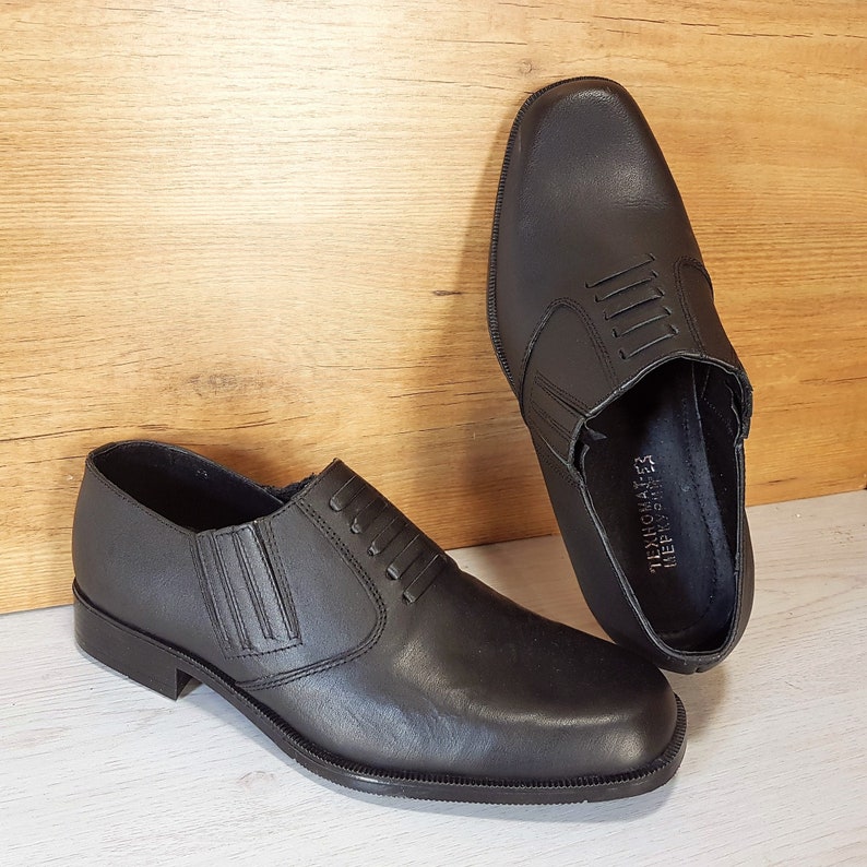 May include: A pair of black leather slip-on shoes with a decorative stitching detail on the vamp. The shoes have a rounded toe and a low heel. The brand name "TECHNOMATES" is printed on the inside of the right shoe.