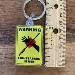 Peut inclure: Un panneau d'avertissement jaune avec un graphique de main noire et de sabre laser. Le panneau indique "ATTENTION SABRES LASER EN UTILISATION". Le panneau est attach&eacute; &agrave; un porte-cl&eacute;s argent&eacute;.