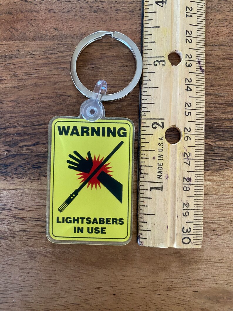 Peut inclure: Un panneau d'avertissement jaune avec un graphique de main noire et de sabre laser. Le panneau indique "ATTENTION SABRES LASER EN UTILISATION". Le panneau est attach&eacute; &agrave; un porte-cl&eacute;s argent&eacute;.
