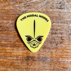 May include: Yellow guitar pick with a black alien face and the text "THE MODAL NODES" printed on it.
