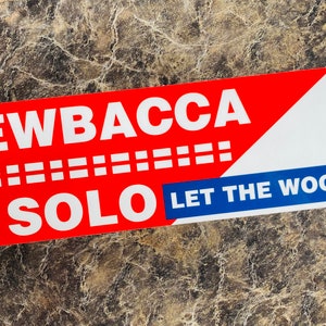 May include: A rectangular sticker with the words "CHEWBACCA SOLO" in white capital letters on a red background. Below, the text "LET THE WOOKIEE WIN" is in blue. The sticker features a stylized American flag design.