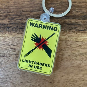 Peut inclure: Un panneau d'avertissement jaune avec un sabre laser noir et une main avec une barre rouge oblique. Le panneau indique "ATTENTION SABRES LASER EN UTILISATION".