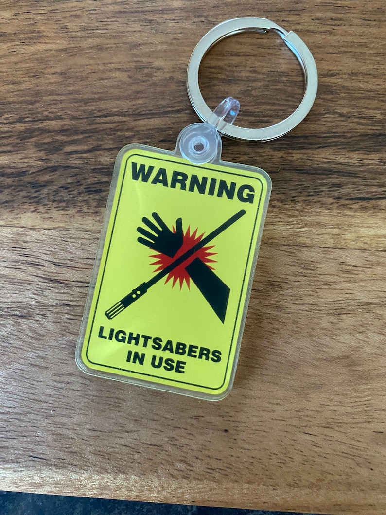 Peut inclure: Un panneau d'avertissement jaune avec un sabre laser noir et une main avec une barre rouge oblique. Le panneau indique "ATTENTION SABRES LASER EN UTILISATION".