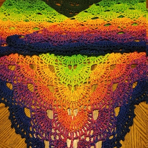 May include: A rainbow crochet shawl with a dark blue border. The shawl is made with a gradient of colors, starting with green and transitioning to yellow, orange, red, purple, and finally dark blue.
