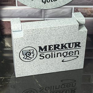 May include: A rectangular, light gray speckled stand featuring the black printed logo and text 'MERKUR Solingen' on the front. The stand has two small, raised rectangular tabs on the top edge, designed to hold a safety razor in an upright position.
