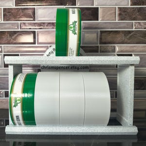 Peut inclure: Un support de rangement gris clair moucheté contenant quatre récipients cylindriques. Trois récipients sont empilés horizontalement sur l'étagère inférieure et un récipient est posé verticalement sur l'étagère supérieure. Les récipients ont des couvercles verts et des étiquettes blanches avec le texte Stirling Soap Co. et Naked & Smooth.