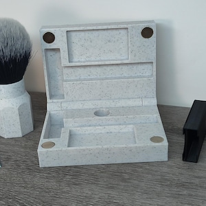 May include: A light gray speckled shaving kit with a hinged lid, compartments, and a black handle. A shaving brush with a white handle and black bristles is to the left. A blue and white Gillette Blue Blade package is visible.
