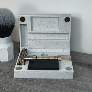 May include: A shaving set featuring a safety razor, a shaving brush, and a case. The razor has a textured handle and is placed on a black base inside the speckled case. A shaving brush with a white and black bristle is also visible.