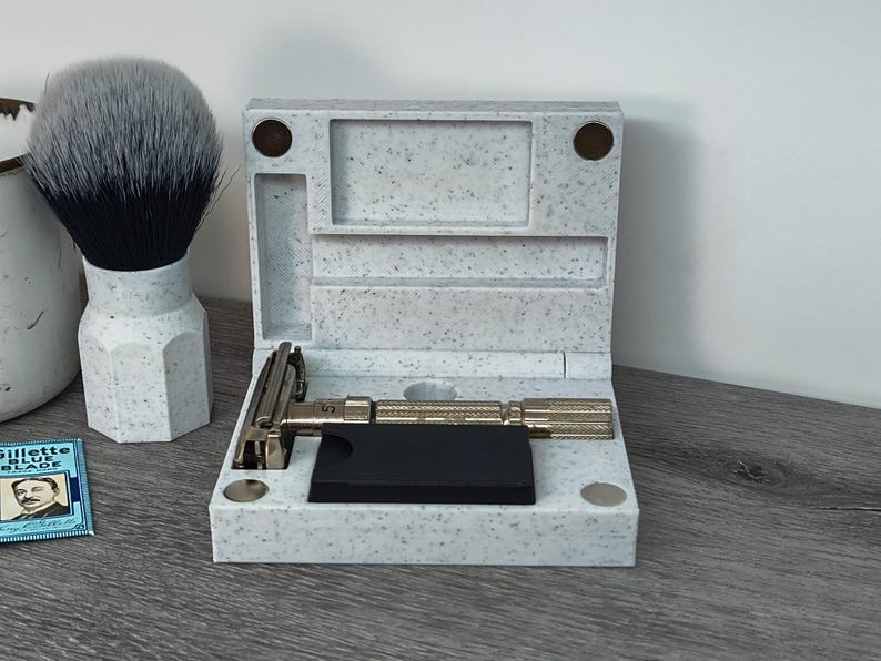 May include: A shaving set featuring a safety razor, a shaving brush, and a case. The razor has a textured handle and is placed on a black base inside the speckled case. A shaving brush with a white and black bristle is also visible.